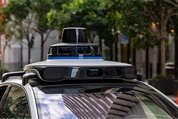 A close-up of a self-driving car’s roof, featuring various sensors and cameras, parked on a city street with trees and buildings in the blurred background.