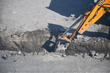 A yellow excavator arm digs a narrow trench in a gray asphalt surface, breaking and lifting the chunks of road. The scene is outdoors and shows construction work in progress.
