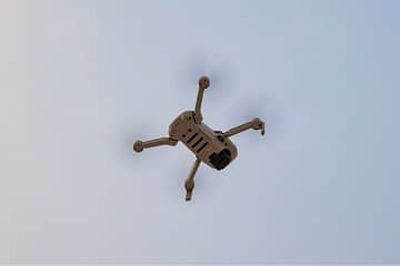 A tan quadcopter drone with four propellers is flying against a clear blue sky, viewed from below. Its camera is visible on the underside of the body.