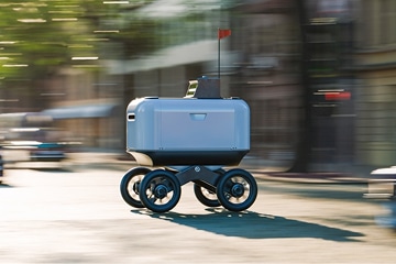 A small, four-wheeled autonomous delivery robot with a red flag on top moves quickly down a city street, with blurred buildings and trees in the background indicating motion.