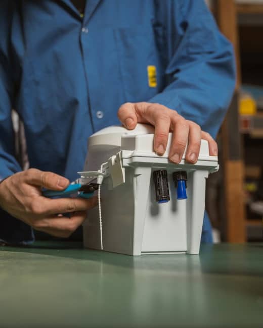 A person in a blue work shirt uses a tool to adjust or assemble a white electrical box with switches and connectors, placed on a green workbench.