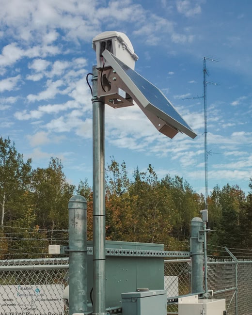 A solar-powered device mounted on a metal pole is seen next to a chain-link fence, with trees and a partly cloudy sky in the background.