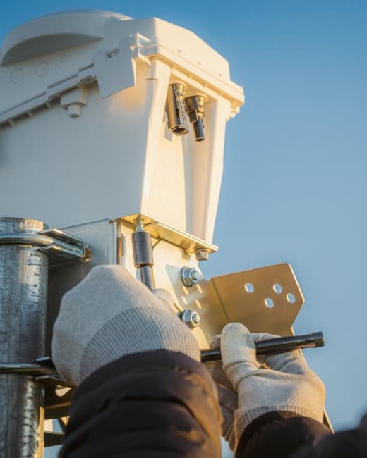 A person wearing gloves installs or adjusts a metal bracket on a telecommunications or electrical equipment box mounted on a pole, with a clear blue sky in the background.