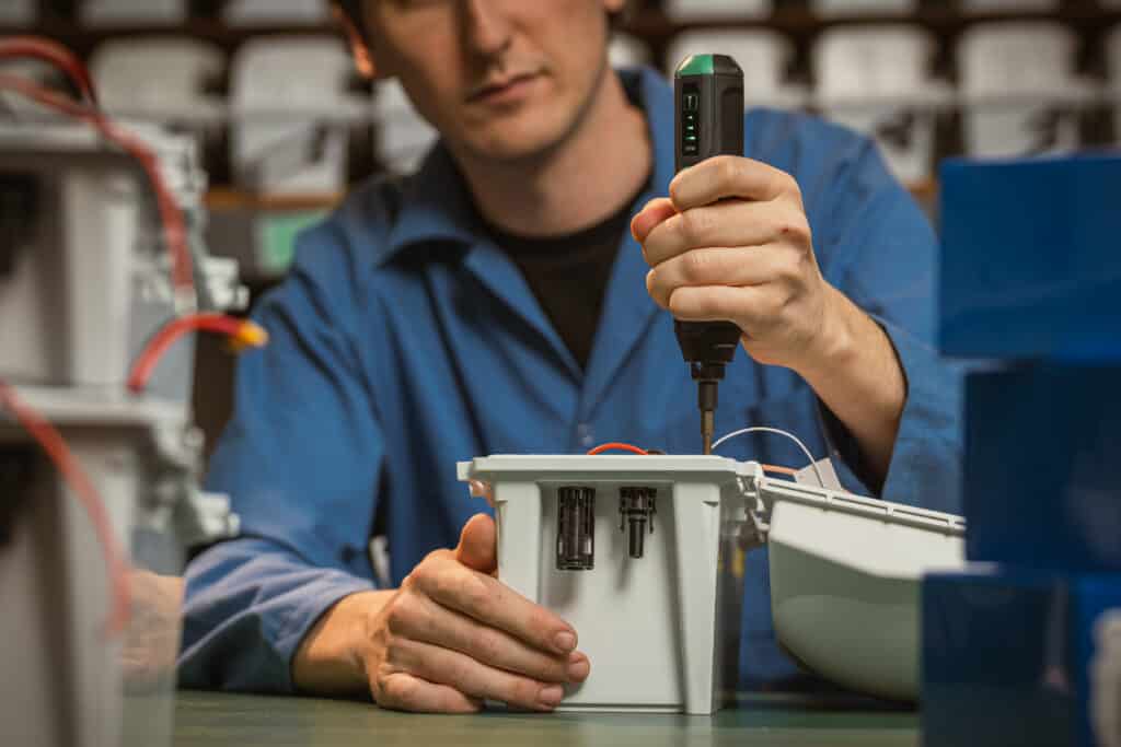 A person in a blue lab coat uses a power screwdriver to assemble an electronic device with exposed wires on a workbench—demonstrating something built, not borrowed.