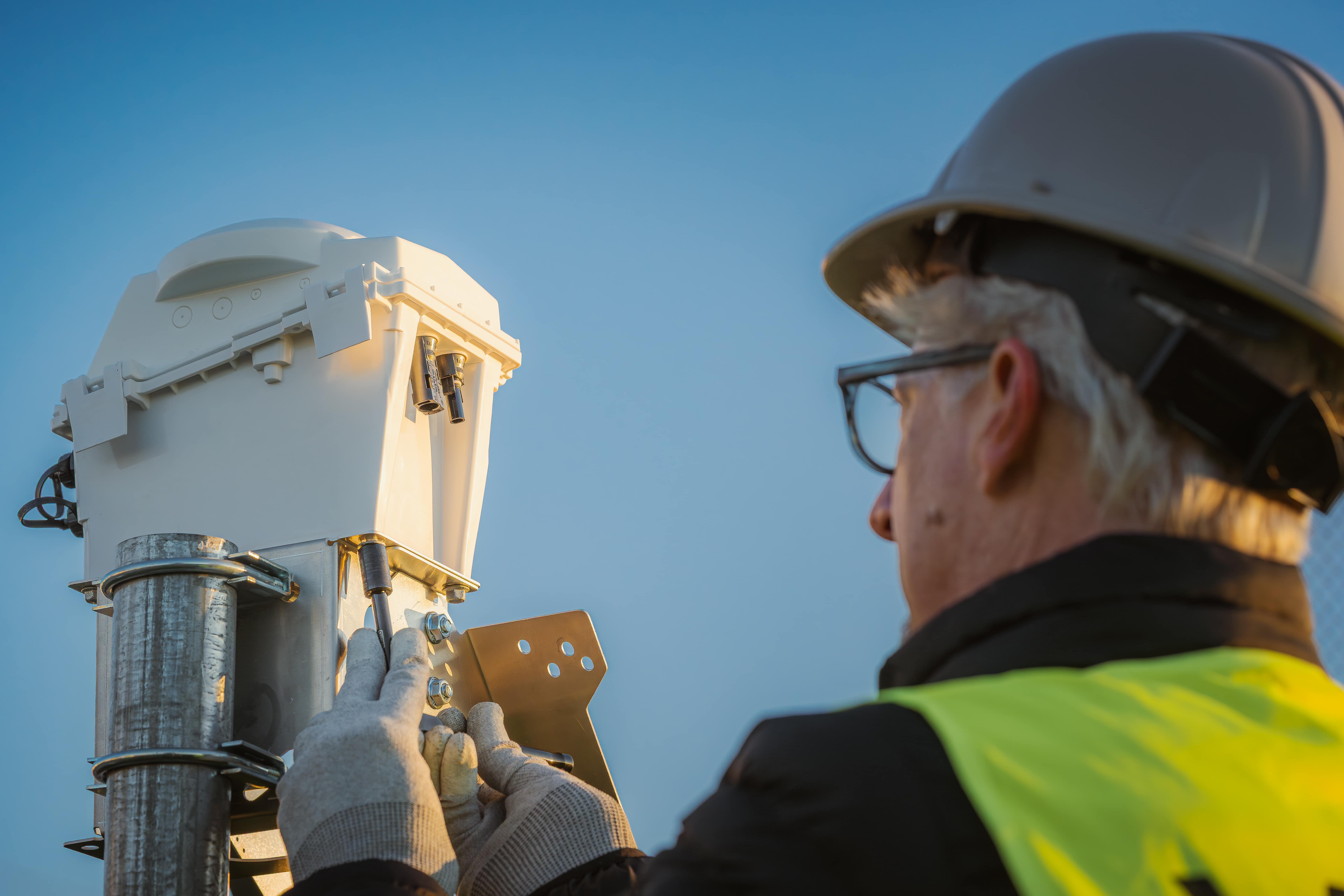 A worker in a hard hat, safety glasses, and a high-visibility vest installs or repairs equipment on a metal pole outdoors under a clear blue sky—demonstrating results that are built, not borrowed.
