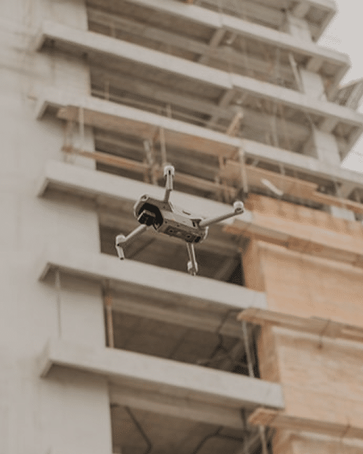 A drone with extended landing gear is flying in front of a partially constructed concrete building with exposed floors and scaffoldings.