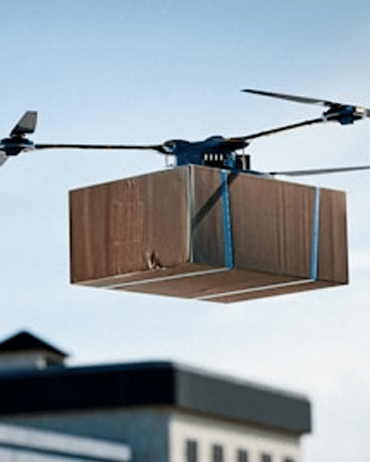 A drone flies in the sky carrying a large cardboard package, with a building visible and slightly out of focus in the background.