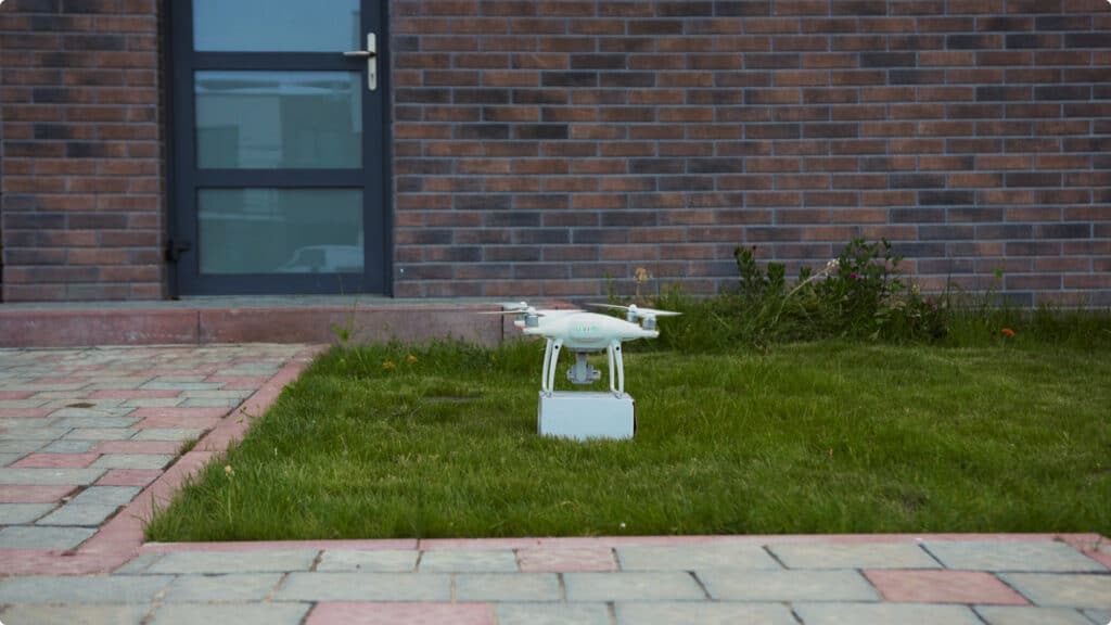 A white drone rests on a small box on a grassy patch in front of a building with a brick wall, glass door, and paved walkway.