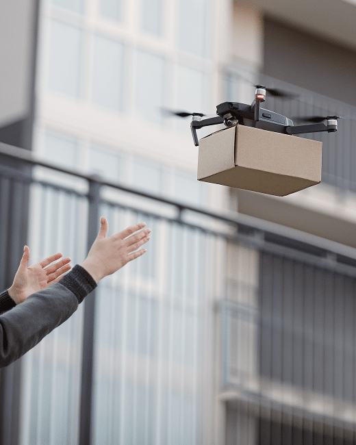 A small drone carrying a cardboard package hovers near a persons outstretched hands, ready for delivery in front of a modern building with glass windows and railings.