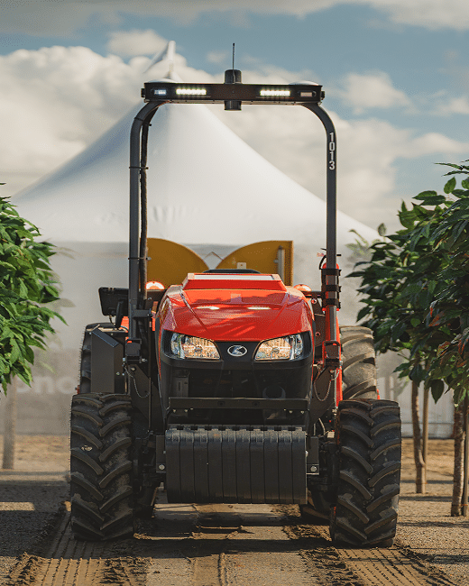 A red tractor with large tires and headlights is parked between rows of green plants, with a white tent and partly cloudy sky in the background.
