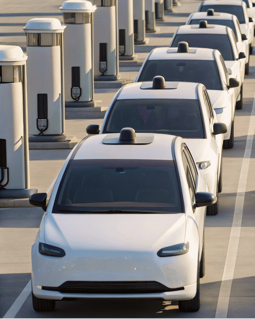 Several white autonomous electric cars are lined up at a charging station, each car connected to a charging unit. The setting appears to be an outdoor parking area under bright daylight.
