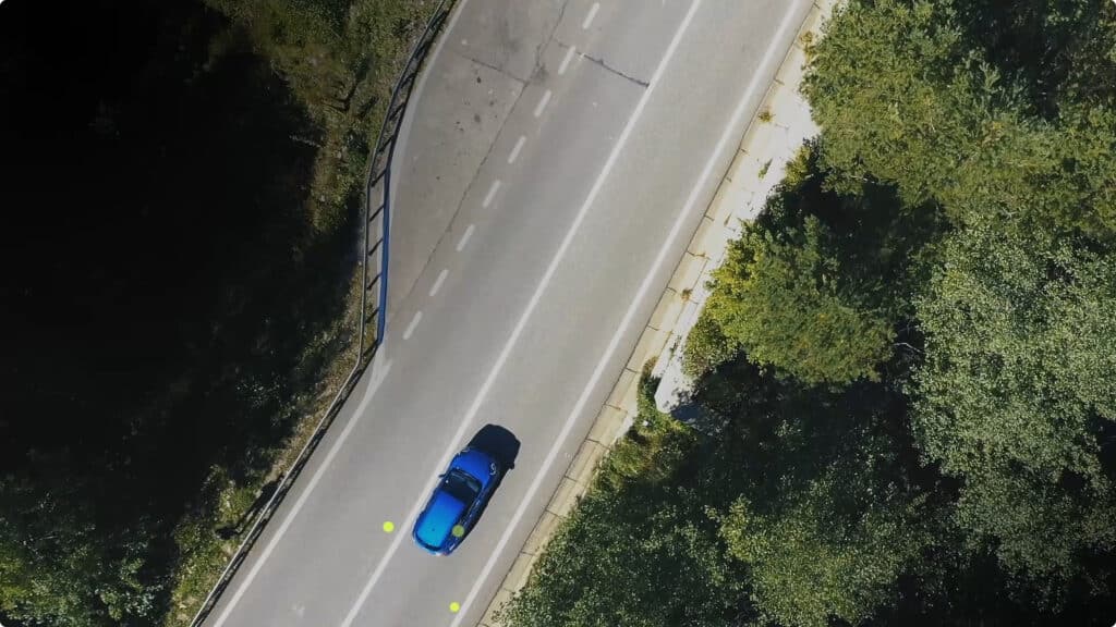 A blue car drives along a two-lane road surrounded by green trees, viewed from above. The road has dashed white lane markings and safety barriers on one side.