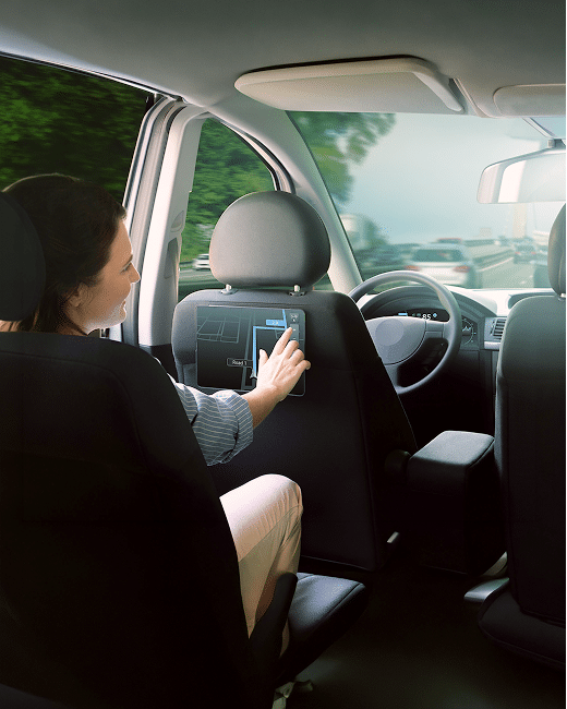 A woman sitting in the backseat of a car touches a screen mounted on the back of the front passenger seat while the car is moving on a road lined with trees.