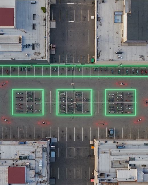 Aerial view of a parking lot between two buildings with three rectangular sections outlined in green neon light, highlighting micro-mobility areas where motorcycles are parked. Empty parking spaces surround the outlined sections.