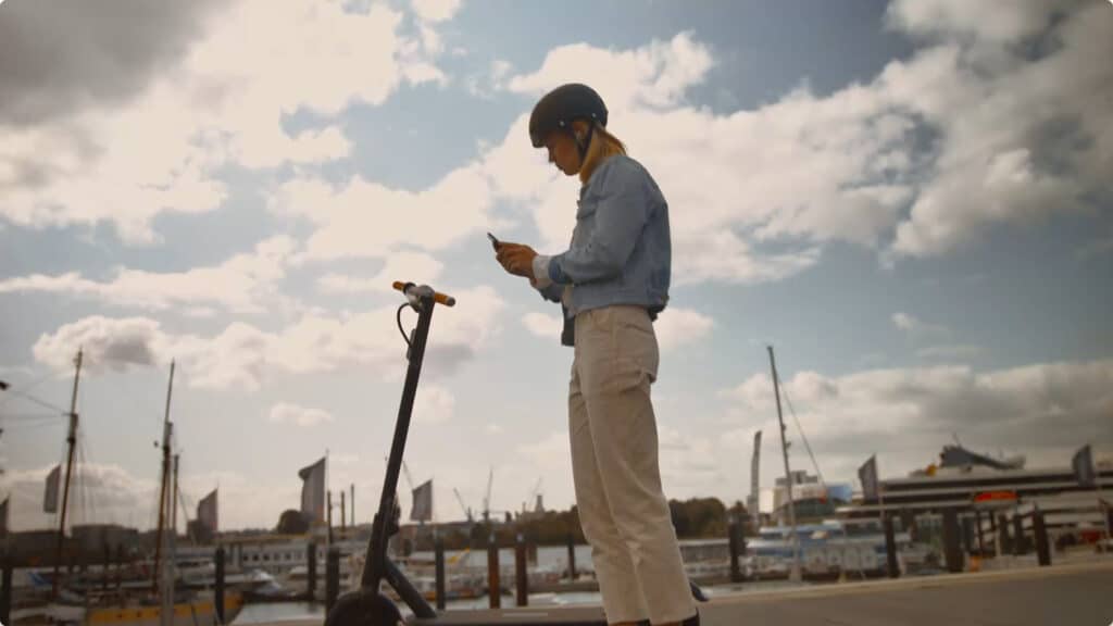 A person wearing a helmet stands outdoors beside an electric scooter, exploring micro-mobility options on a smartphone. The background features a harbor with boats and a partly cloudy sky.