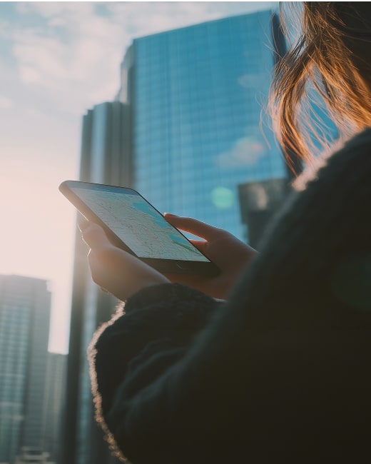 A person holding a smartphone and looking at a map app, standing outdoors in a city with tall glass buildings in the background, illuminated by sunlight.