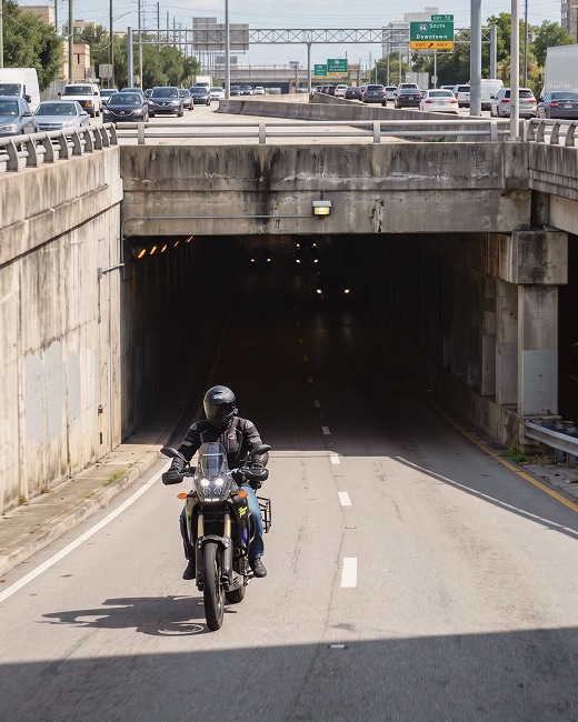 A motorcyclist wearing a helmet and black gear rides out of a tunnel onto a sunlit road, with heavy traffic and cars visible on the overpass above.
