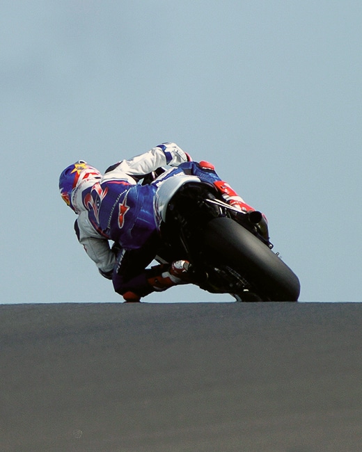 A motorcycle racer leans sharply into a turn on a racetrack, wearing a purple and white racing suit and helmet, with the rear tire close to the ground against a clear sky.
