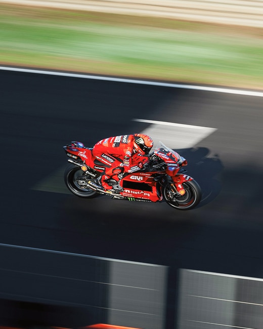 A motorcycle racer in red gear and helmet rides a red racing motorcycle at high speed on a track, with motion blur in the background.