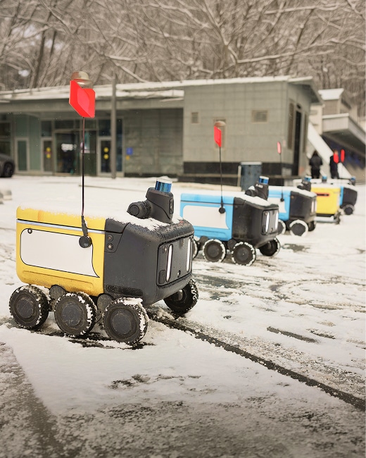 Three small, boxy delivery robots with six wheels each, two blue and one yellow, lined up in the snow. Each has a red flag on top and is in front of a modern building with trees in the background.