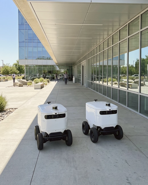 Two white delivery robots with wheels are driving on a sidewalk outside a modern glass building. A few people are walking in the background along the covered walkway.