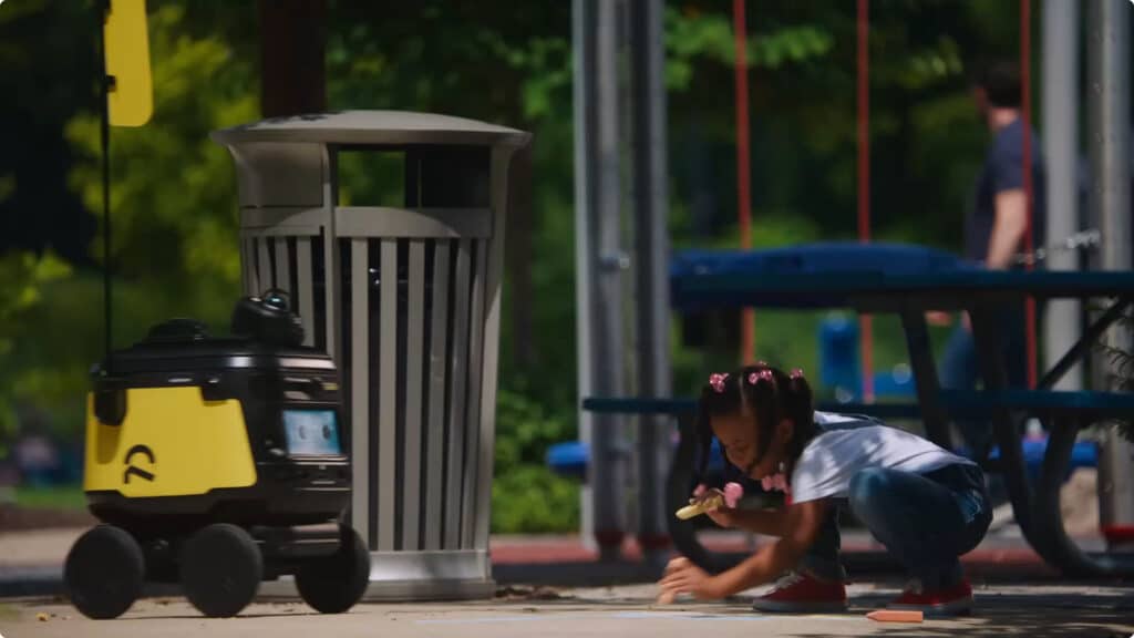 A young girl crouches on a sidewalk drawing with chalk near a trash can, while a small delivery robot with a yellow flag rolls by in a park setting.