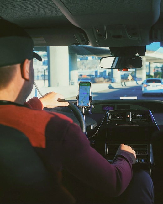 A person driving a car is looking at a smartphone mounted on the dashboard, displaying a navigation map, while sunlight streams in through the windshield.