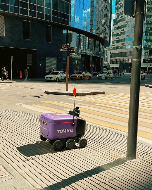 A small purple delivery robot with wheels and a red flag is on a sidewalk near a pedestrian crossing in a modern city with glass buildings and parked cars.