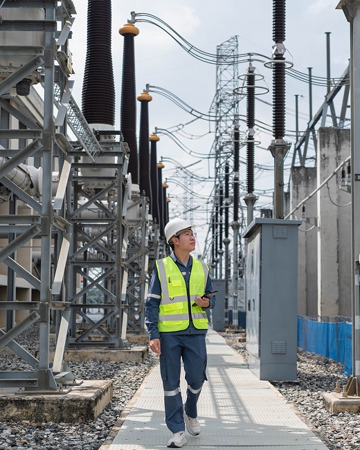 A worker in a safety vest and hard hat walks through an outdoor electrical substation, surrounded by large power equipment and high-voltage towers.