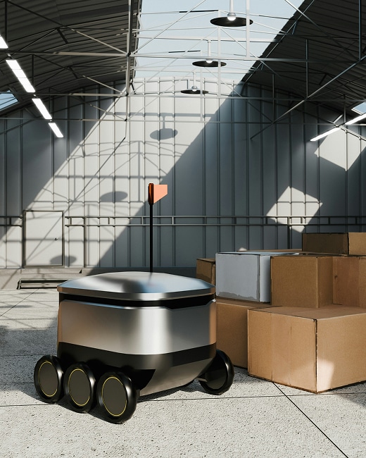 A small, sleek delivery robot with six wheels and an orange flag is positioned next to cardboard boxes inside a spacious, modern warehouse with metal walls and a glass ceiling.