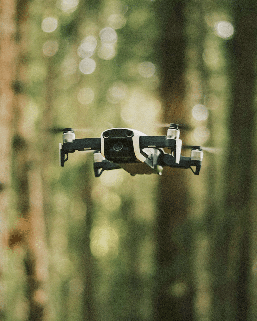 A black drone with four propellers hovers in mid-air, surrounded by tall, blurred trees in a sunlit forest. The background is softly focused, highlighting the drone in the foreground.