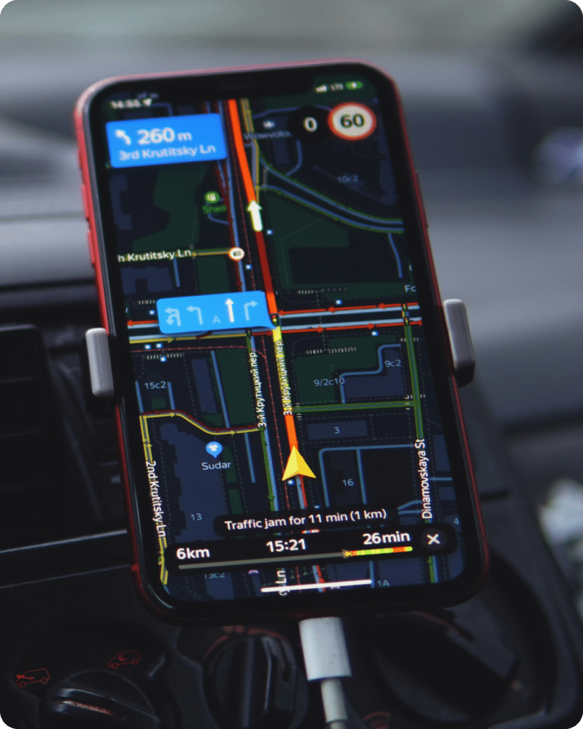 A smartphone mounted in a car displays a GPS navigation app with directions, showing a route, traffic information, and upcoming turns on a map. The device is charging via a cable.