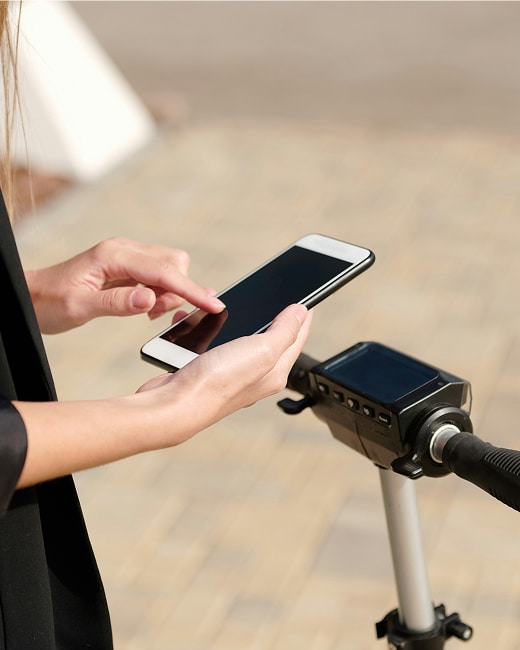 A person using a smartphone while standing next to a micro-mobility electric scooter, with one hand holding the phone and the other touching the screen. The scooter’s handlebar and display are clearly visible.