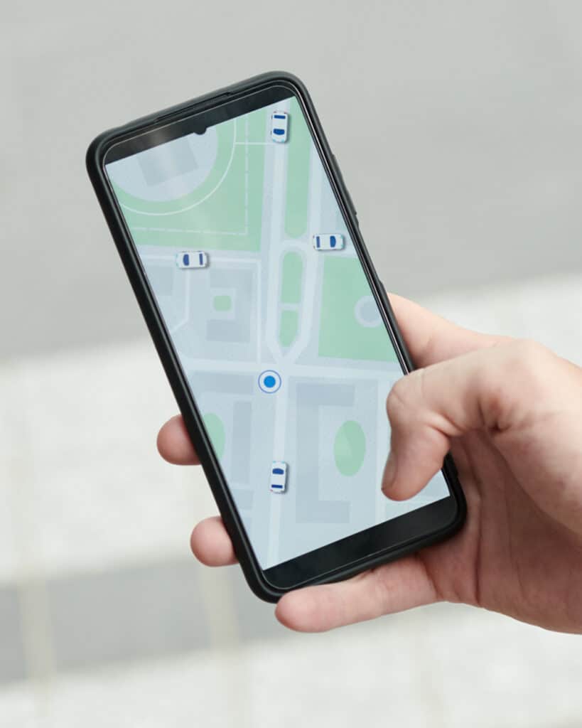 A hand holding a smartphone displaying a map with several car icons, indicating nearby vehicles, likely on a ride-hailing or navigation app.