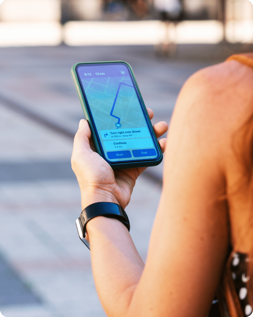 A person holding a smartphone with a navigation app open, showing directions and a route on a map. The person is wearing a black wristwatch and standing outdoors.