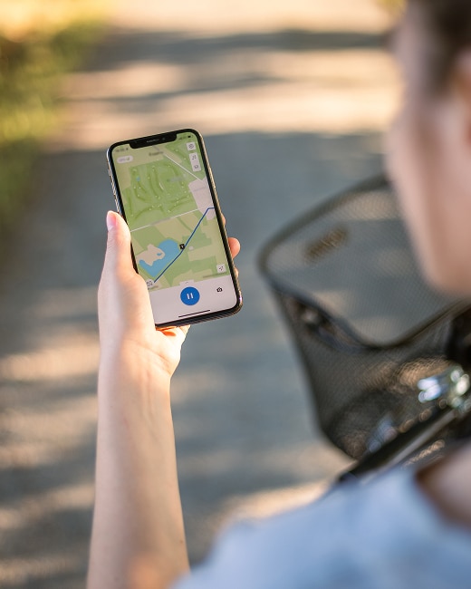 A person holds a smartphone displaying a navigation map while riding a bicycle outdoors, with a bike basket and a sunlit path visible in the background.