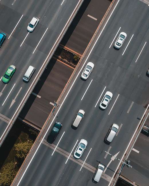 Aerial view of multiple lanes of a highway with several cars traveling in both directions, showing clear roads, white lane markings, and some greenery visible between the road sections.