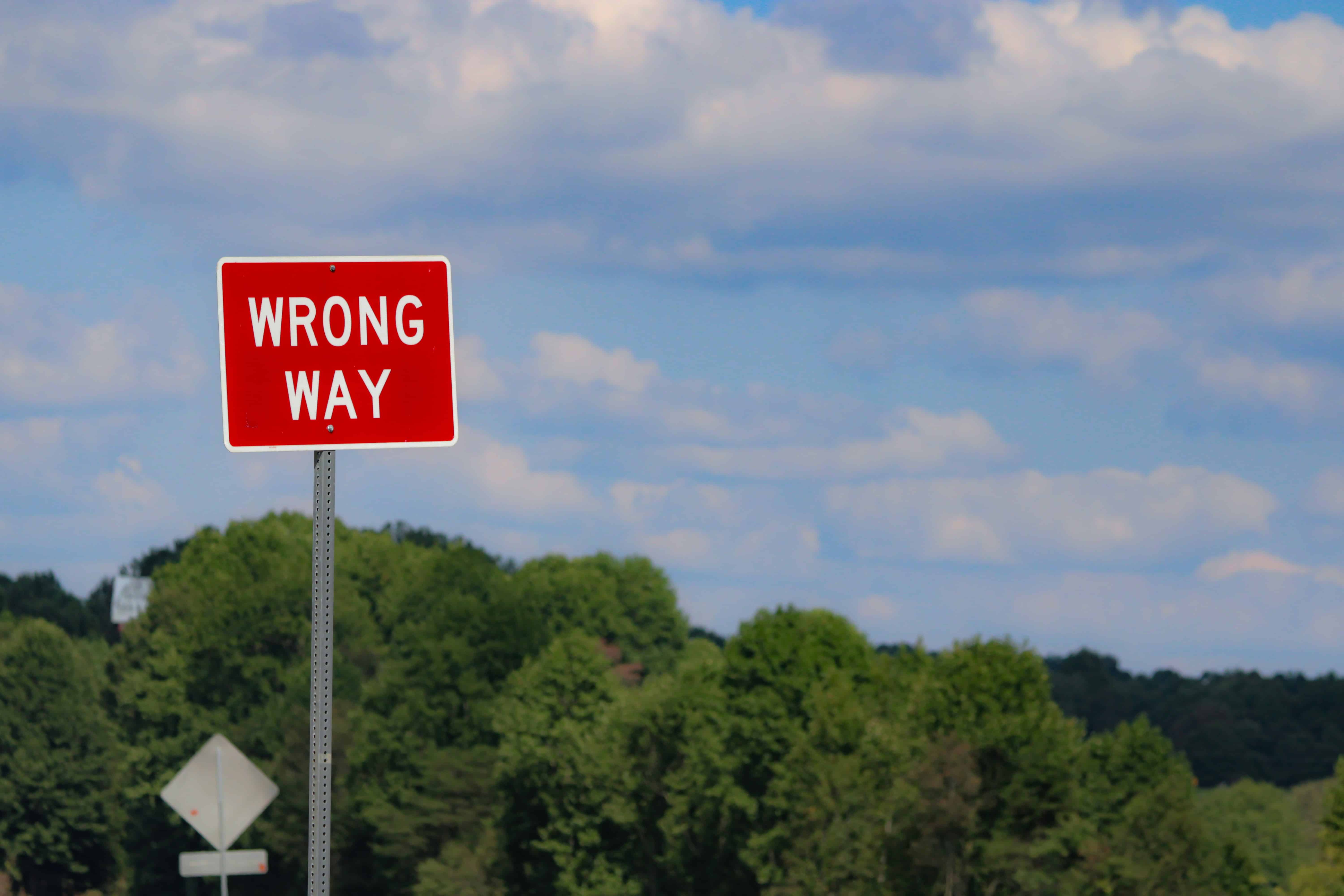 A red WRONG WAY road sign stands in front of green trees, with a blue sky and scattered clouds in the background.