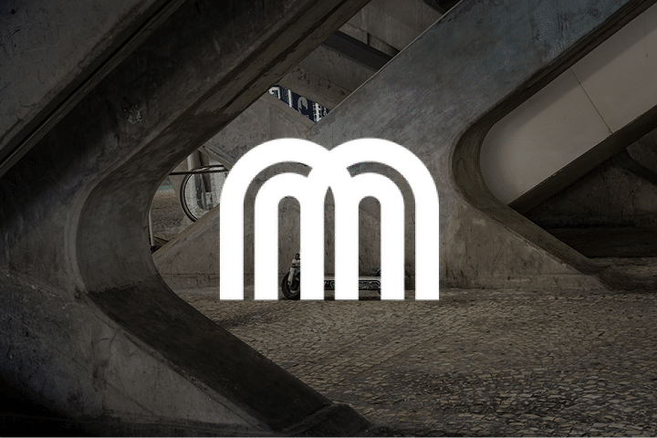 A white metro logo with three arches is centered over a background of concrete beams and a tiled floor, possibly inside a subway or train station, evoking the innovative spirit found at events like the Micromobility America conference.
